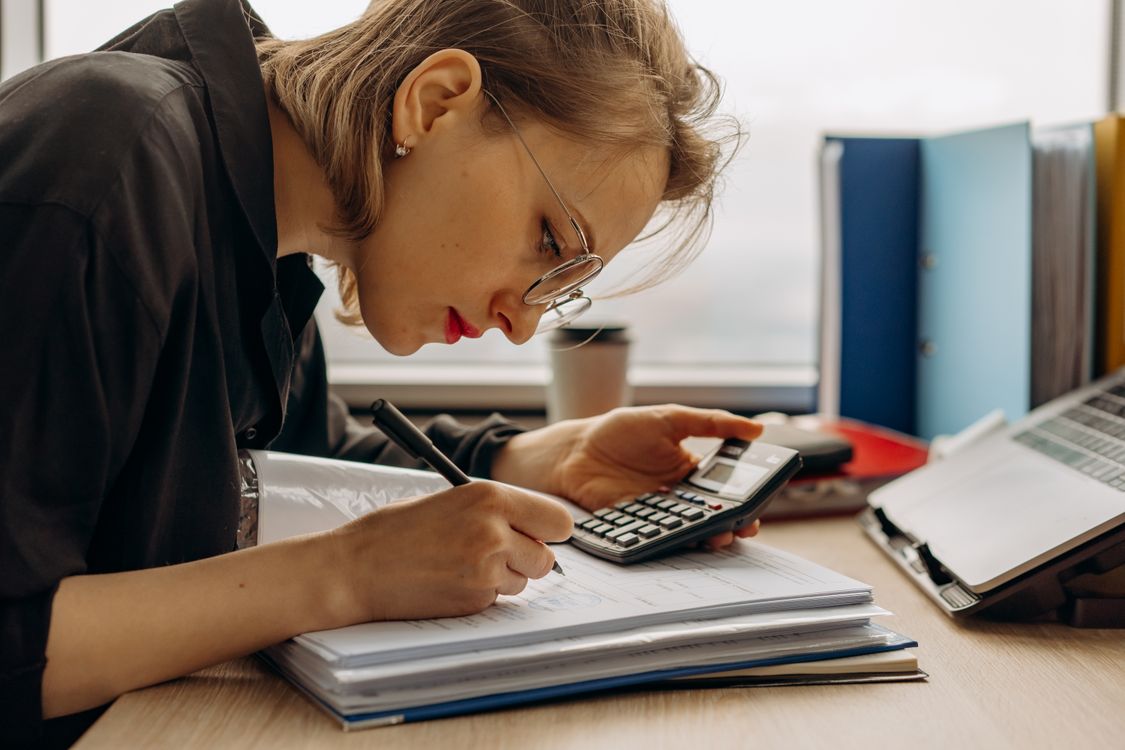 A young woman works on accounting at a desk, writing in documents while holding a calculator beside a laptop.
