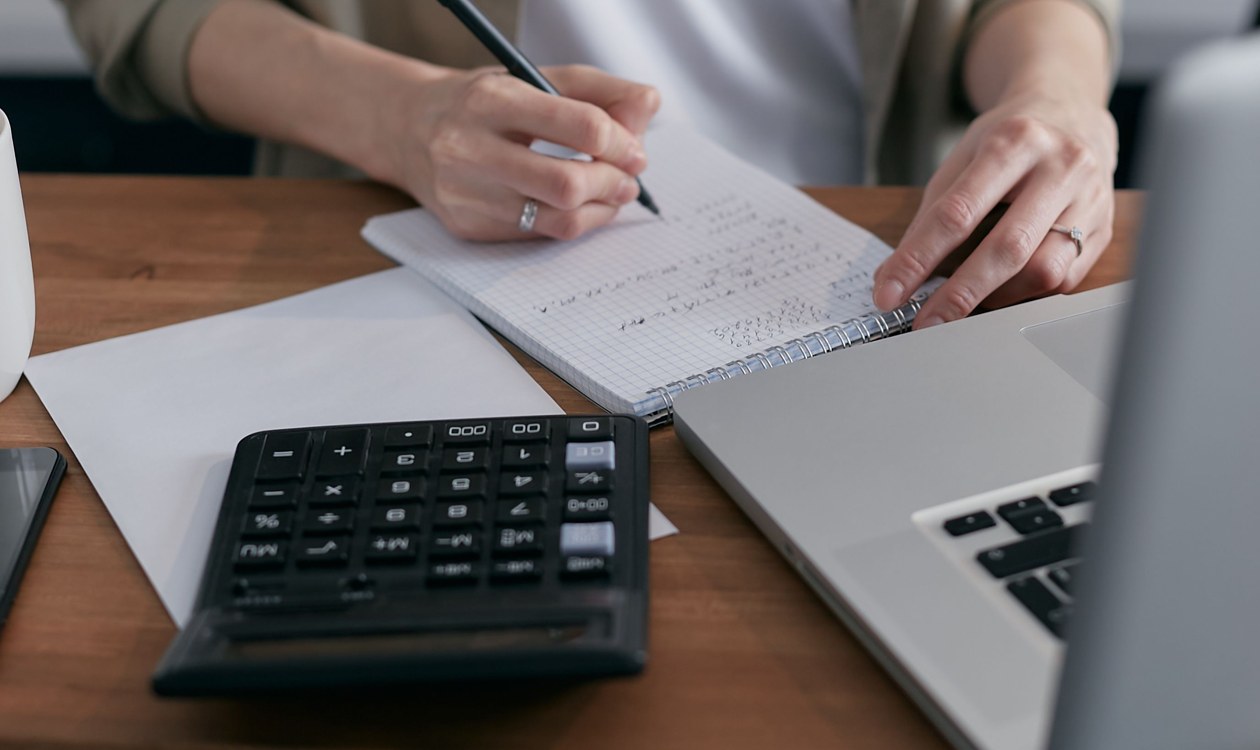 Woman working through figures in a notebook at her desk with a calculator and laptop